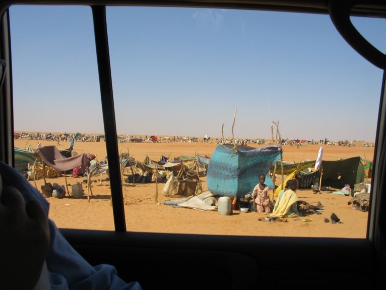 Figuur 1 Foto zichtbaar genomen vanuit het zijraam van een auto. Te zien is hoe mensen in een woestijnachtige omgeving, in de zon, onder een lichtblauwe hemel, kleine schuilplaatsen hebben gemaakt met stokken, doeken en plastic.
